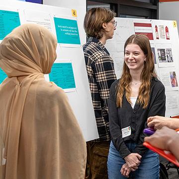 在STEM海报研讨会上，一名年轻女子礼貌地向一群聚集在她海报周围的人微笑